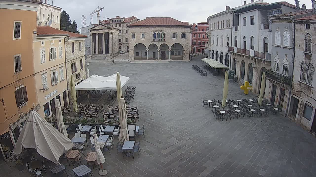A large, mostly empty town square with several outdoor cafe seating areas under umbrellas, surrounded by stone buildings on an overcast day.