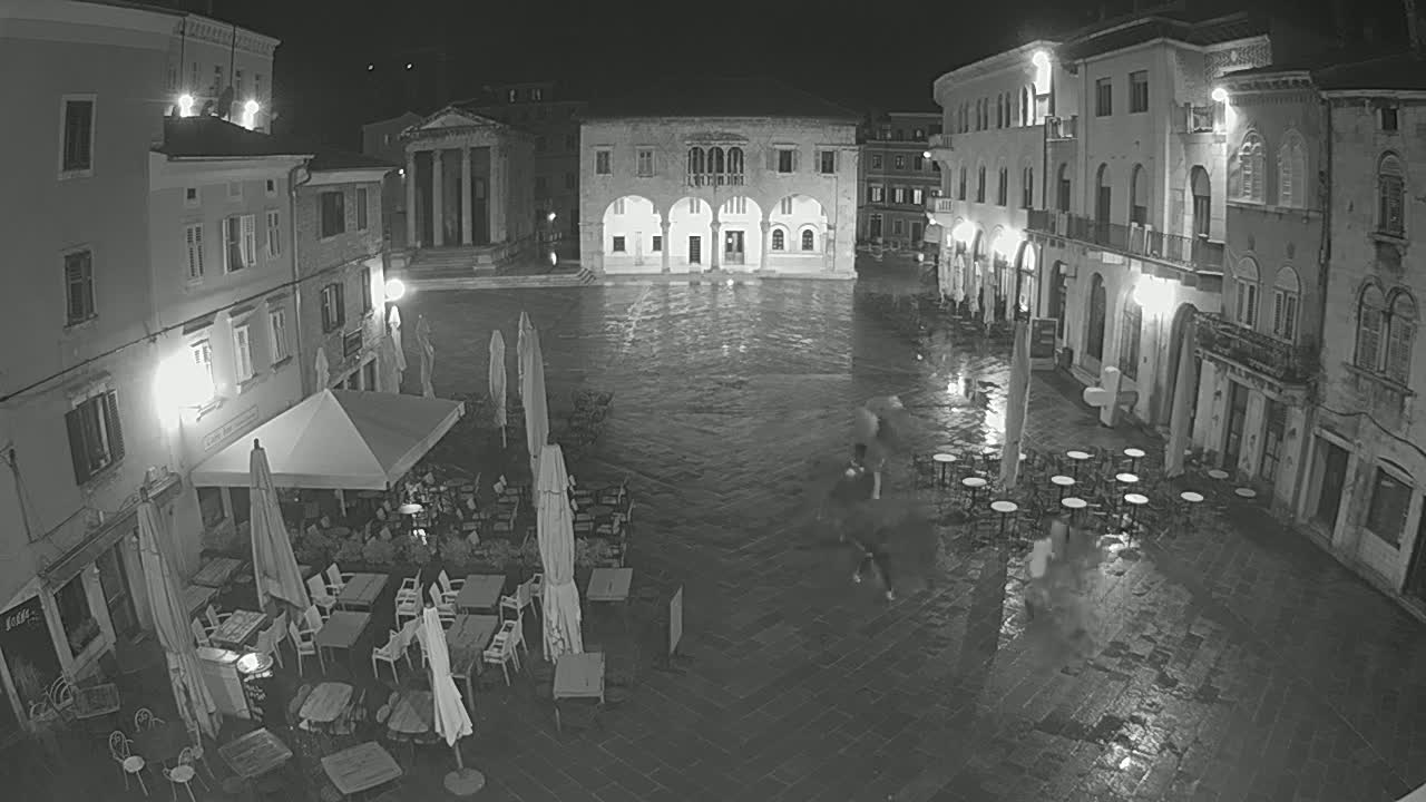 A rain-slicked, nighttime town square is sparsely populated with several people and outdoor cafe seating under umbrellas.
