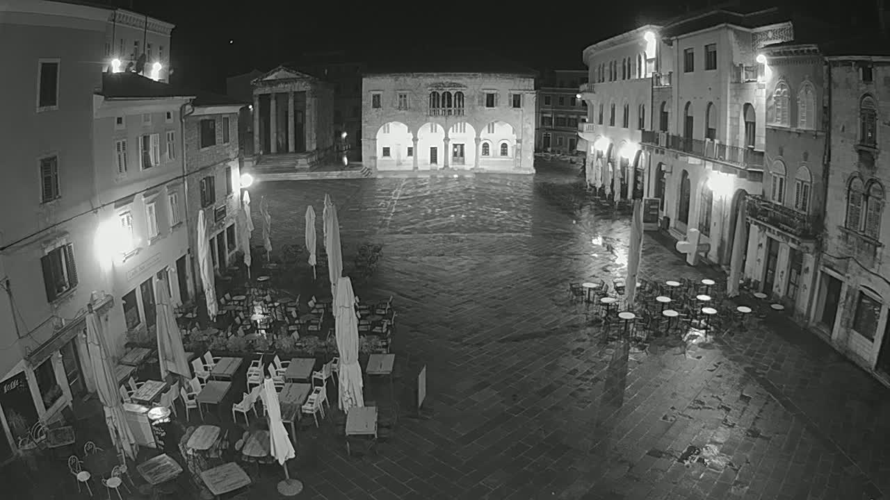 A wet, empty town square at night, surrounded by old buildings with outdoor café seating under umbrellas.