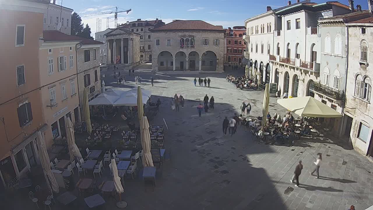 A town square with several outdoor cafes and pedestrians under a sunny sky.