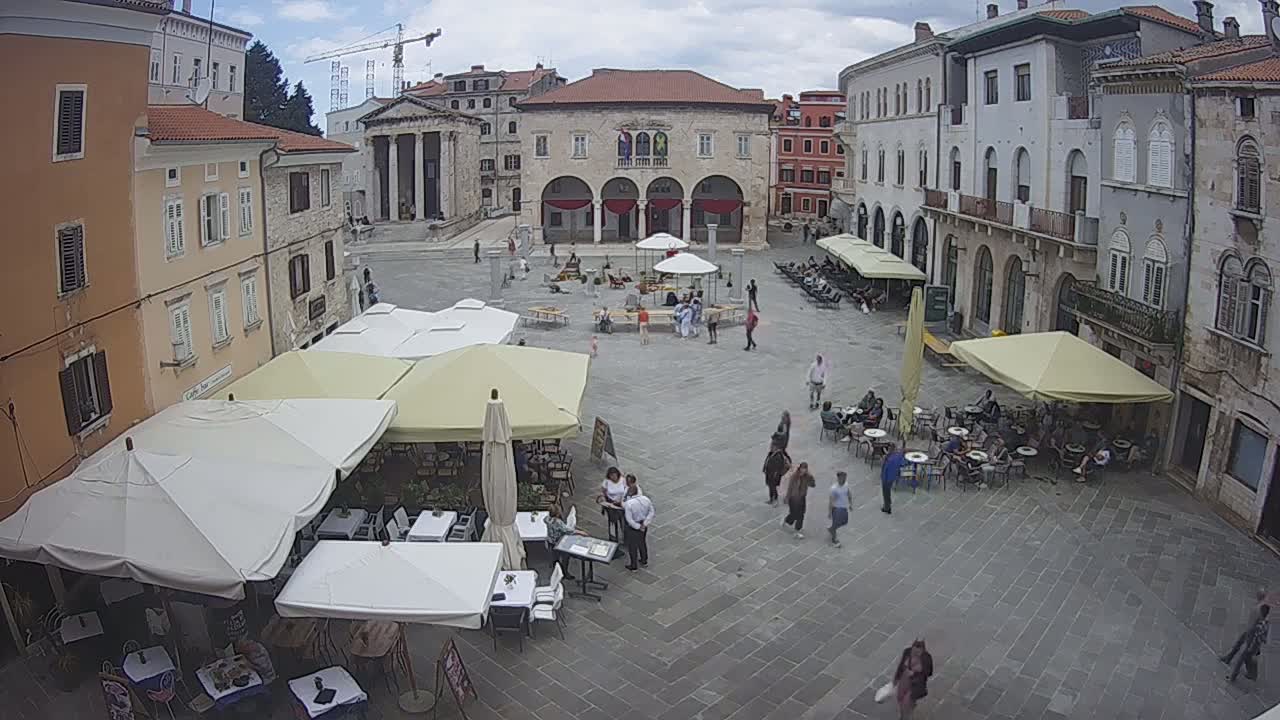 A town square with several outdoor cafes under umbrellas, surrounded by buildings under a partly cloudy sky.