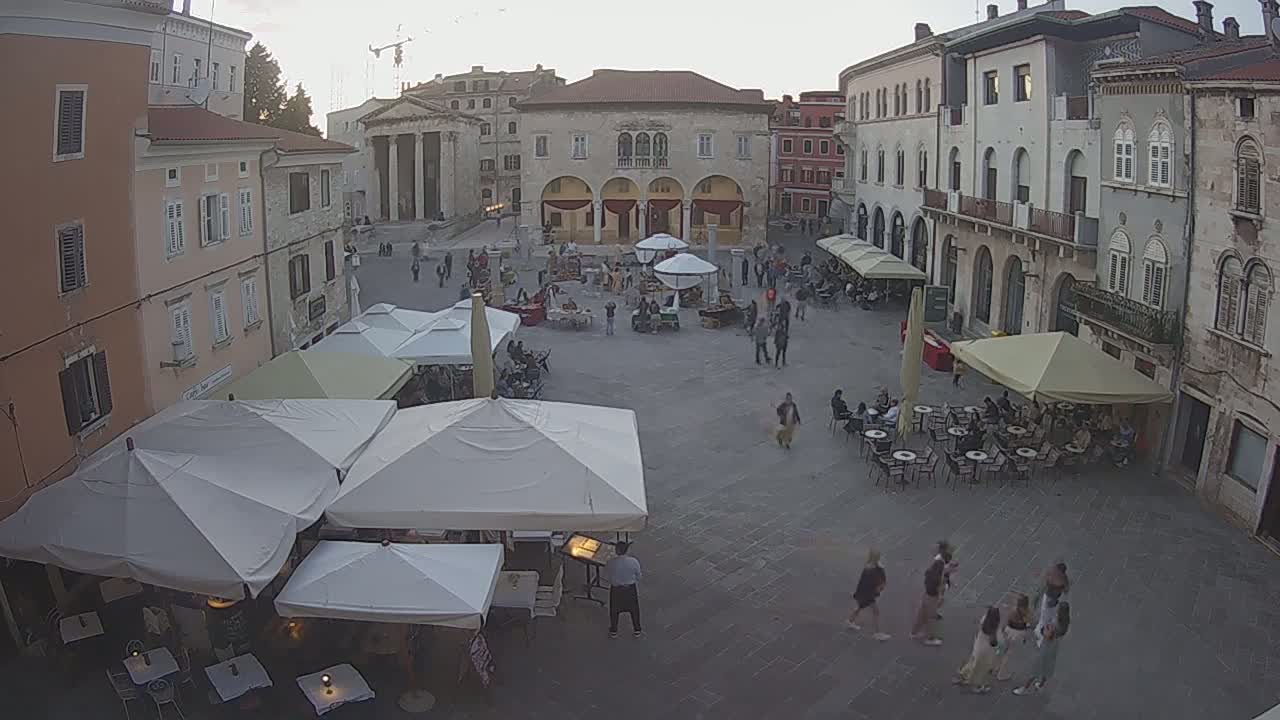 A town square, under a partly cloudy evening sky, is filled with people sitting at outdoor cafes and strolling around market stalls beneath white umbrellas.