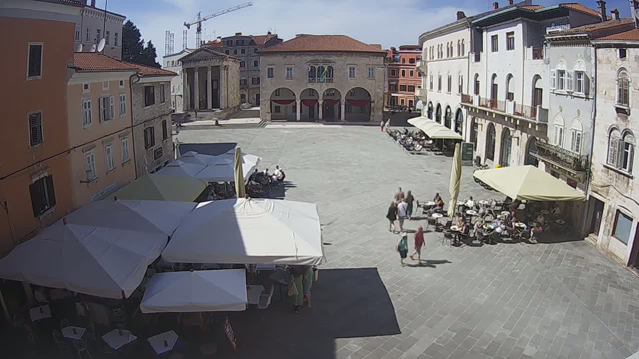 A sunny day in a large European-style town square with several outdoor cafes under umbrellas and people walking around historic buildings.