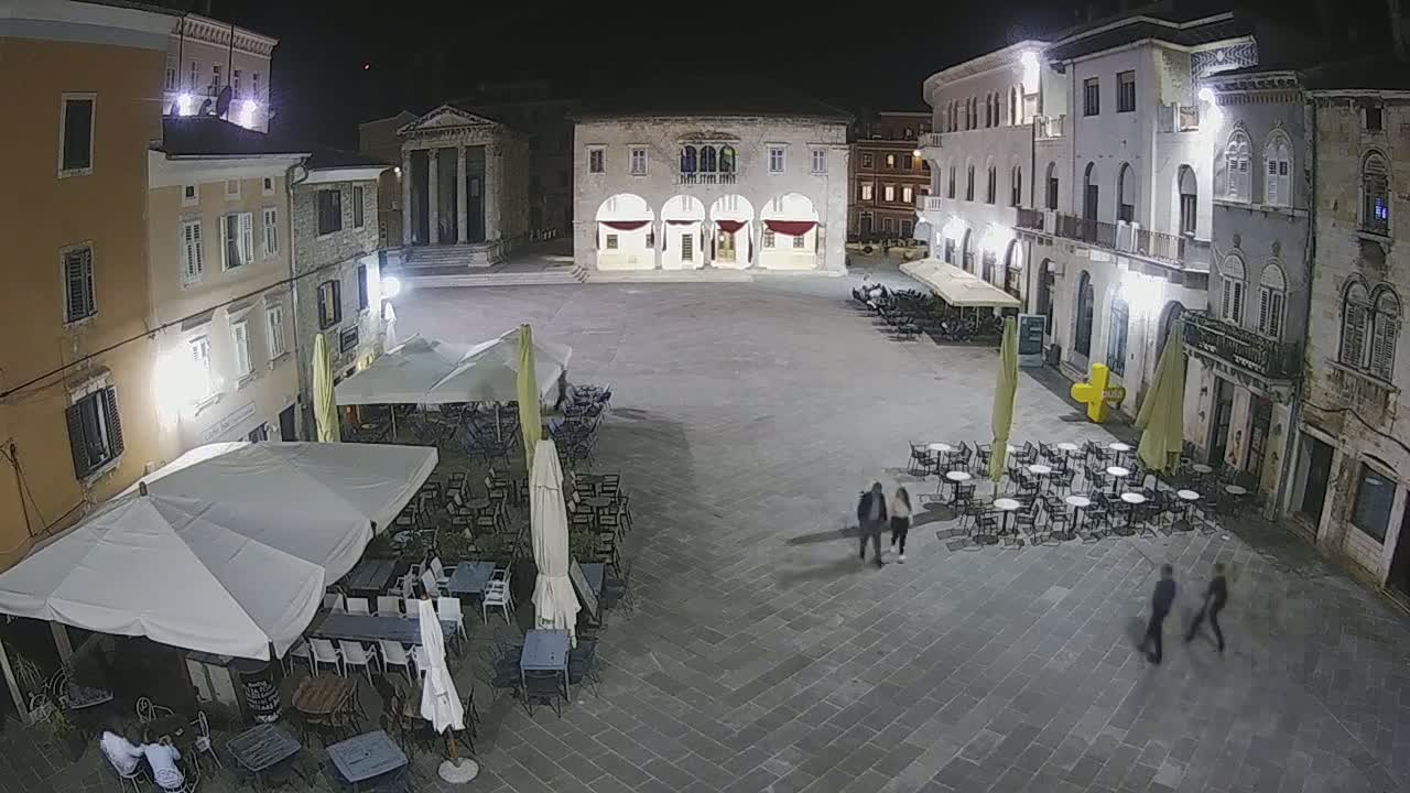 A nighttime view of a town square with several outdoor cafes under umbrellas, surrounded by stone buildings.