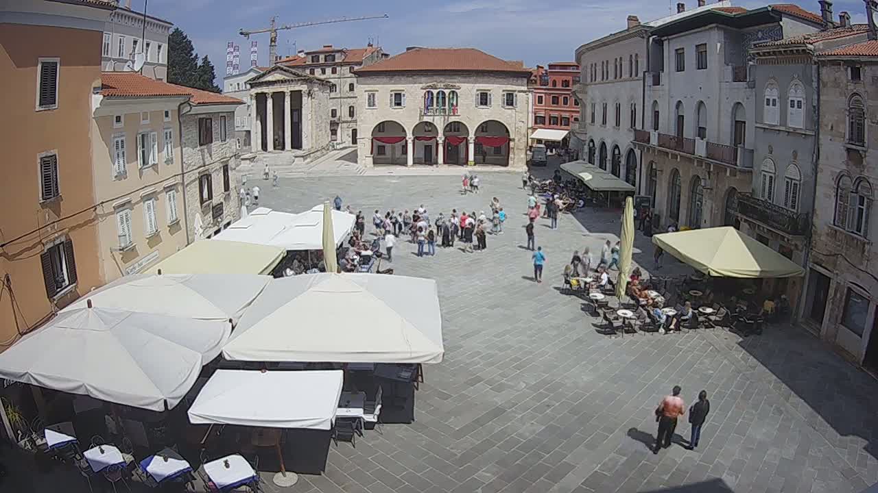 A sunny town square with many people gathered and seated at outdoor cafes under umbrellas.