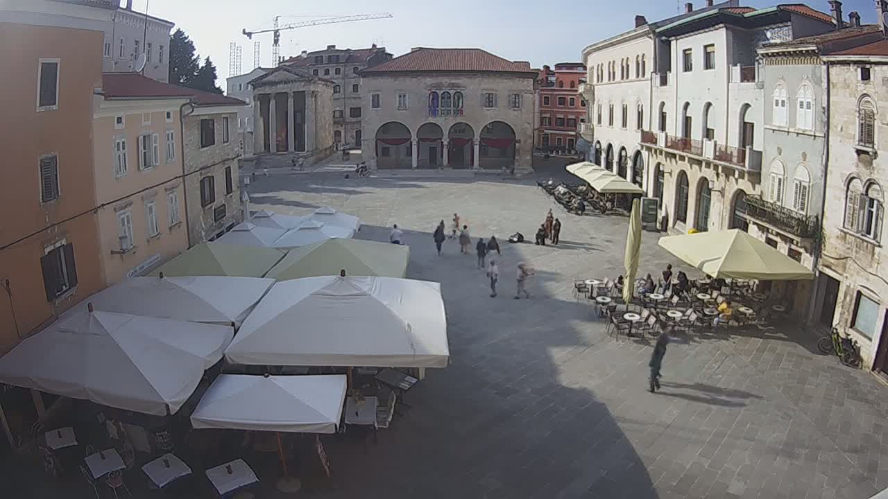A sunny town square with several outdoor cafes under white and beige umbrellas, surrounded by historical buildings.
