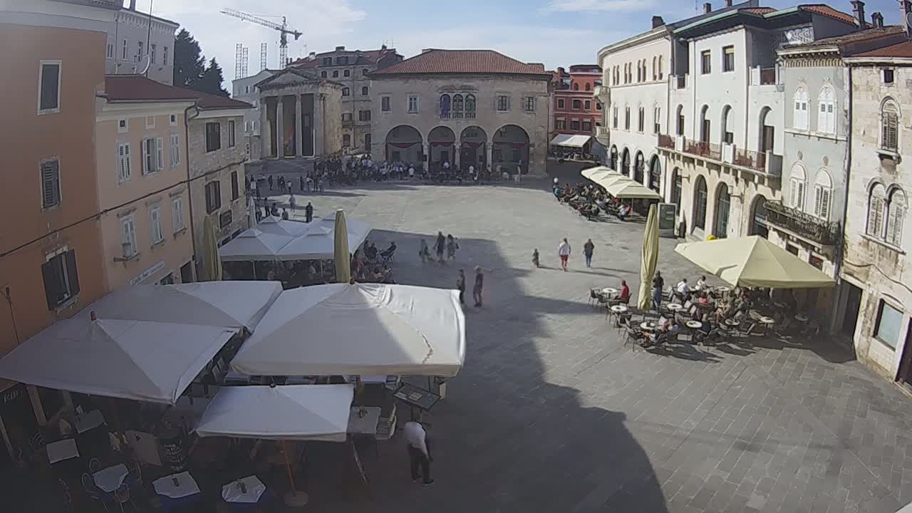 A large town square with several outdoor cafes under umbrellas, surrounded by old buildings on a sunny day.