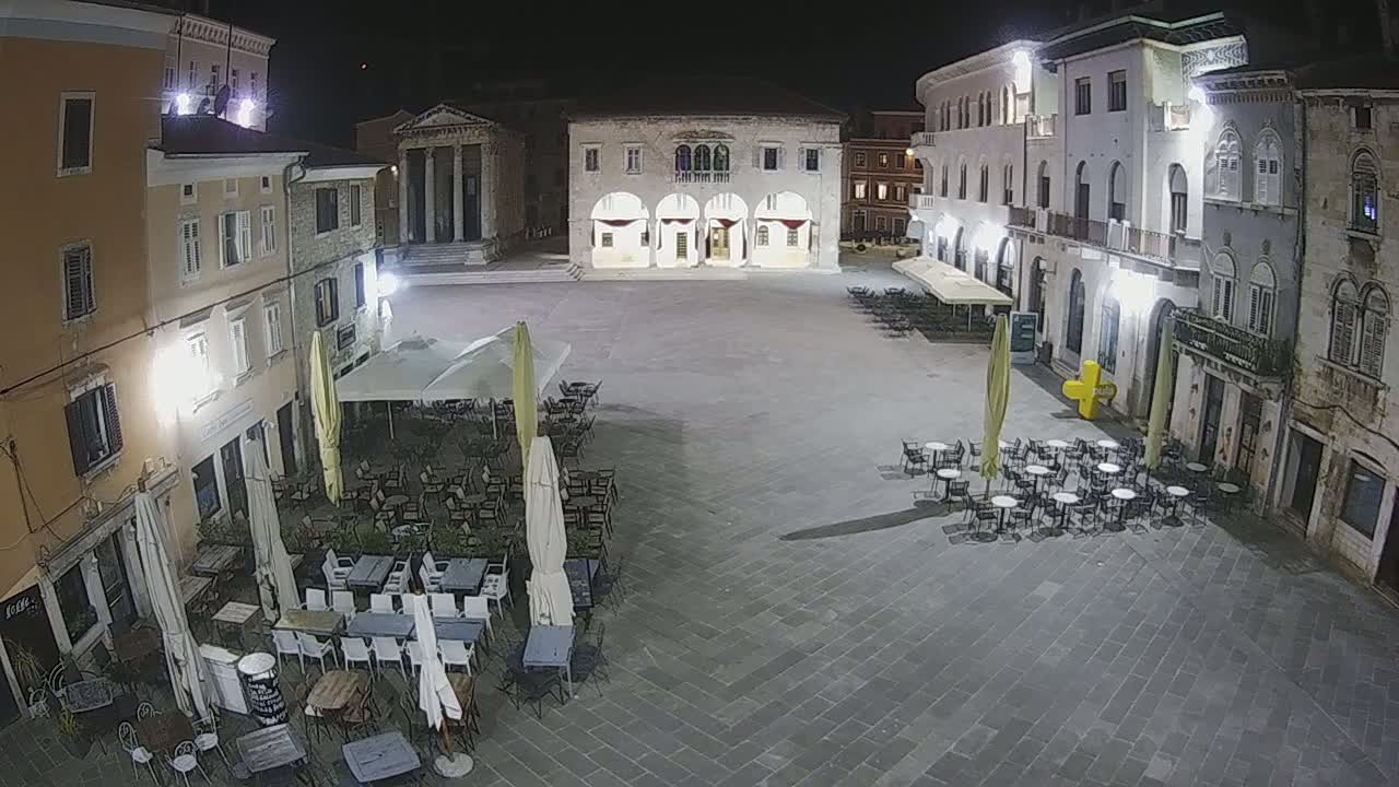 A nighttime view of a town square with several outdoor cafes and old buildings surrounding it under well-lit conditions.