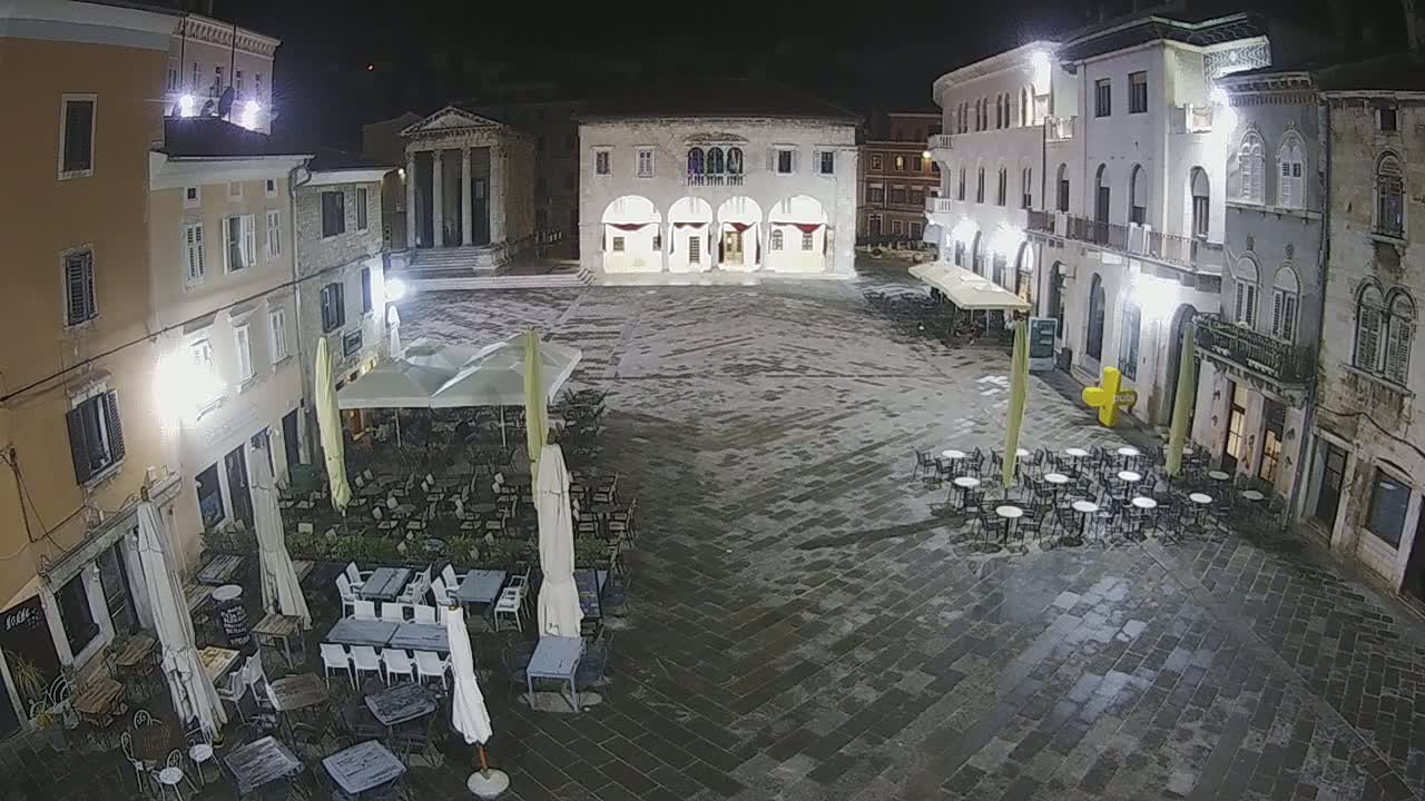 A nighttime view of a town square with several buildings surrounding it, outdoor seating at cafes under umbrellas, and a slightly wet cobblestone pavement.