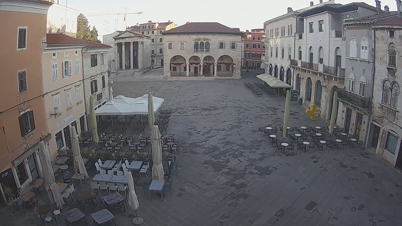 A large, mostly empty town square with several outdoor cafe tables and umbrellas under a clear morning sky, surrounded by old stone buildings.