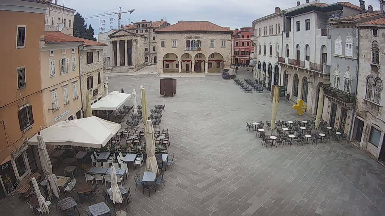 An overcast day reveals a town square with numerous outdoor café tables and chairs arranged around a large, stone building, surrounded by other buildings with visible architectural details.