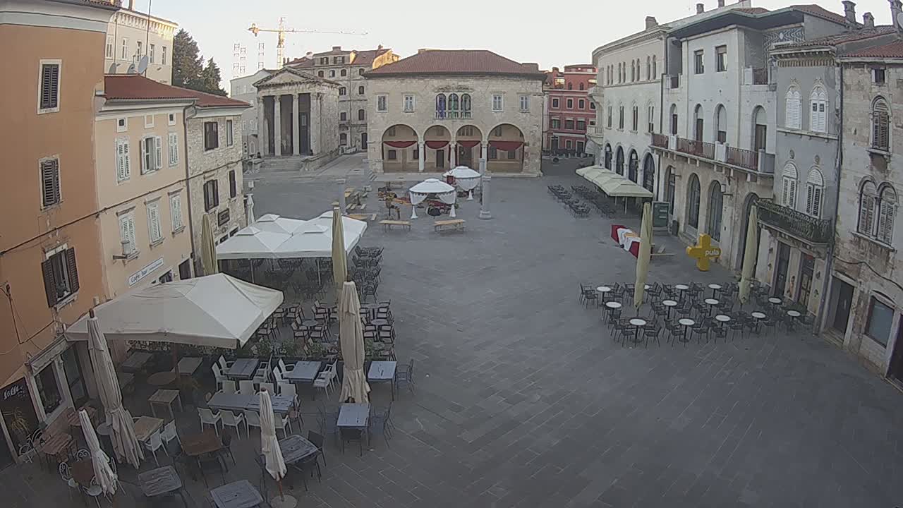 A mostly empty town square with several outdoor cafes under umbrellas, surrounded by old stone buildings under a clear sky.