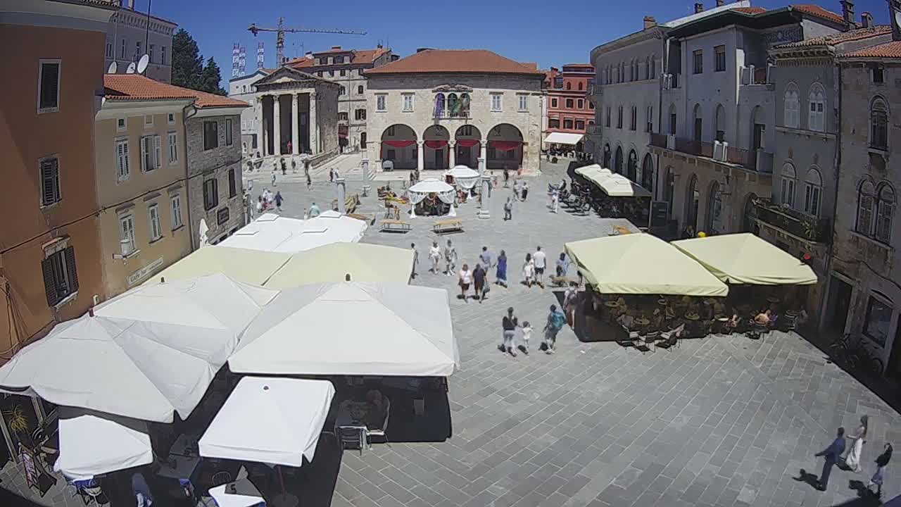 A sunny day in a European town square shows many people strolling among several outdoor cafes with patio umbrellas.