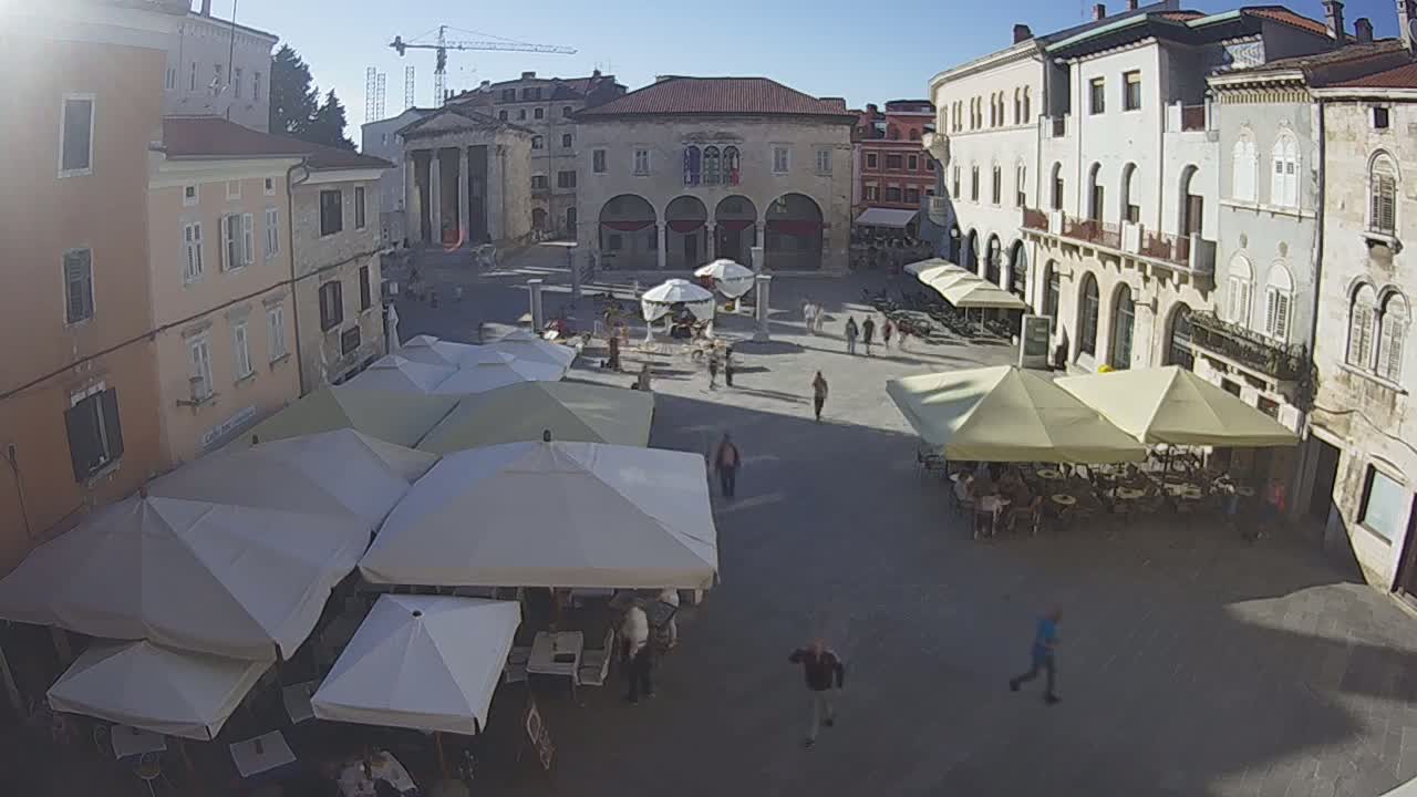 A town square with several outdoor cafes under umbrellas, surrounded by multi-story buildings on a sunny day.