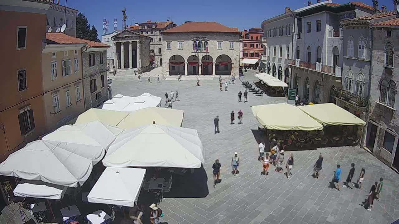 A town square with several outdoor cafes under shade umbrellas, surrounded by historic buildings on a sunny day.