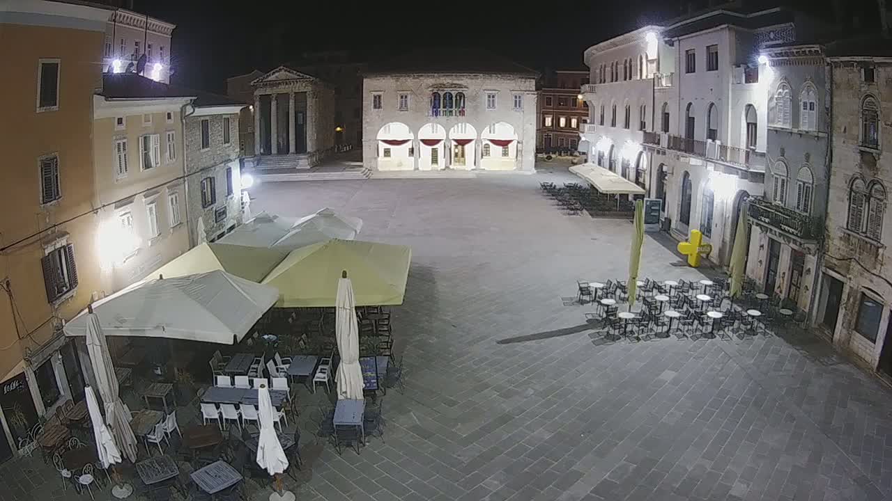 A nighttime view of a town square with several outdoor café umbrellas and tables under artificial light.