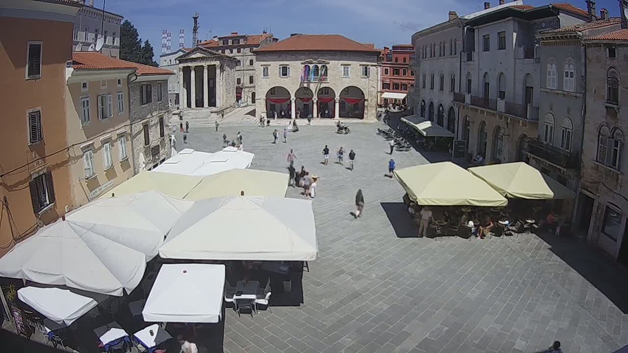 A sunny day in a large town square with several outdoor cafes under umbrellas, surrounded by historic-looking buildings.