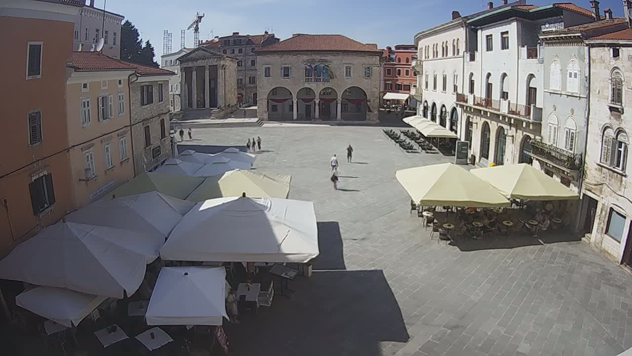 A sunny town square with several outdoor cafes under parasols, surrounded by old stone buildings.