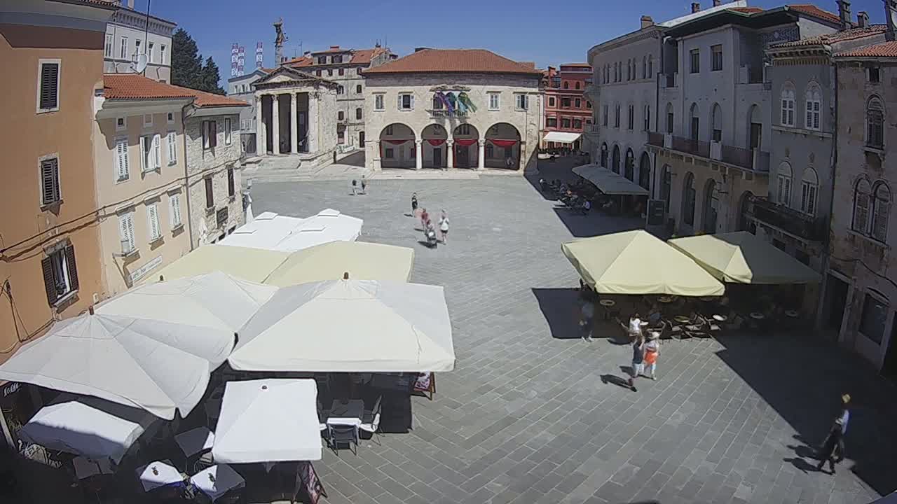 A sunny town square with several outdoor cafes under umbrellas, surrounded by old stone buildings.