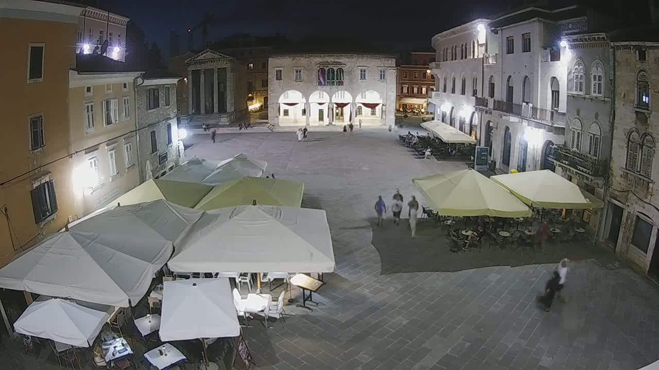 A nighttime, high-angle view of a town square with several outdoor cafe umbrellas set up around the perimeter, and a few people walking around.