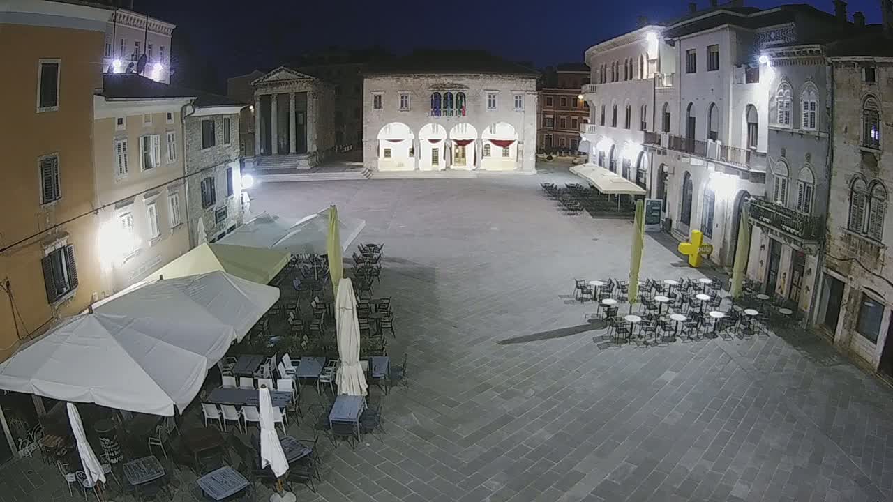 A town square at night, illuminated by streetlights, with several outdoor cafe tables and umbrellas under a clear, dark sky.