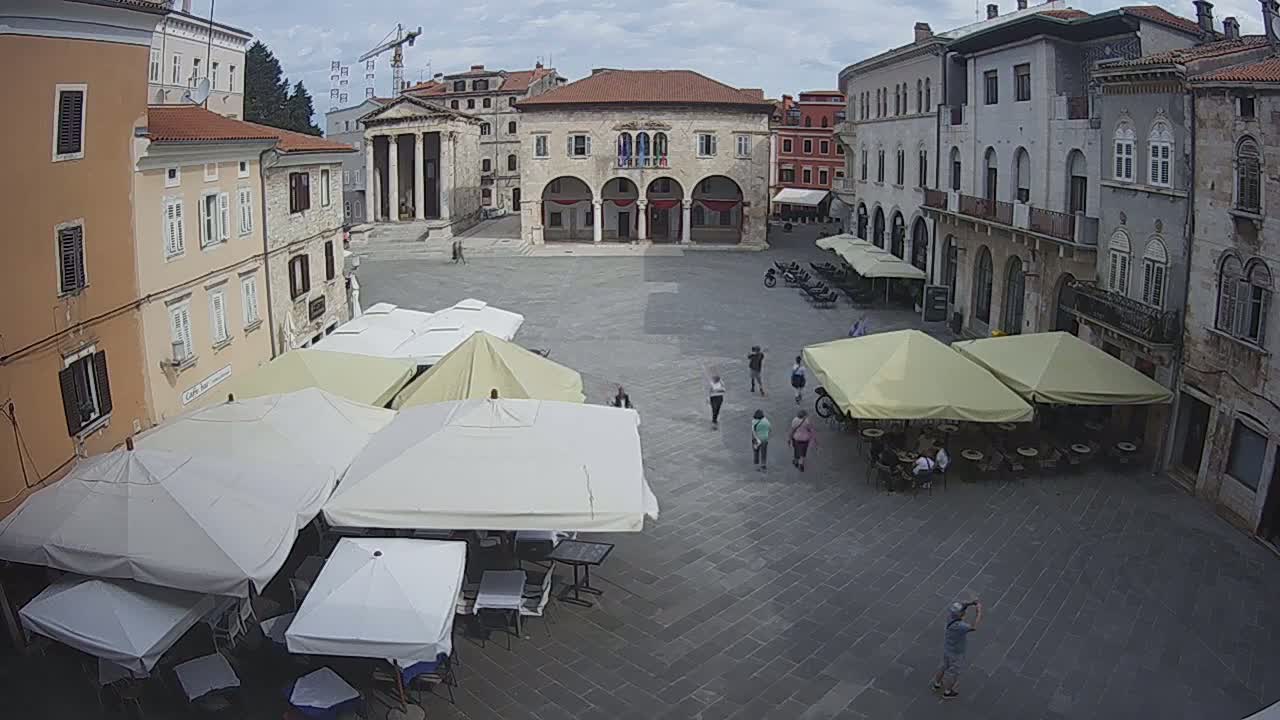 A large town square with several outdoor cafes under umbrellas, surrounded by historical buildings on a partly cloudy day.