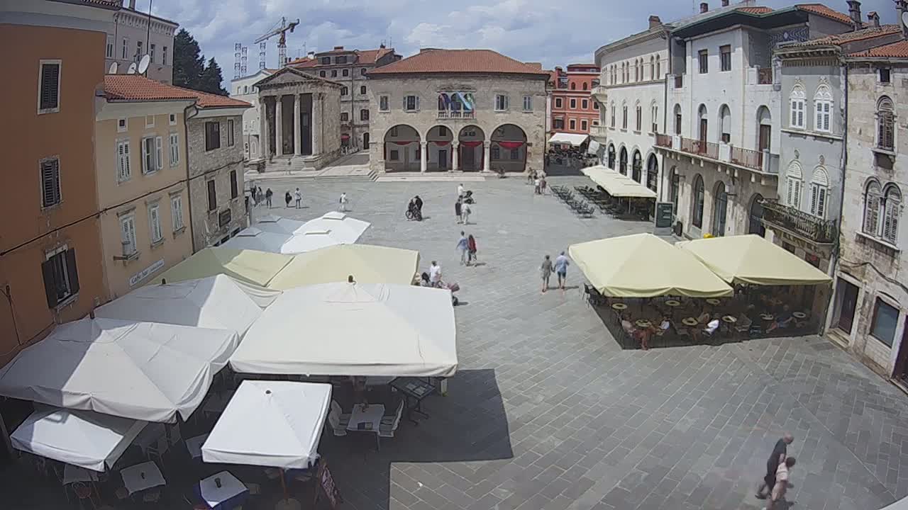 A paved town square with several people walking around and several cafe umbrellas set up under mostly sunny skies.
