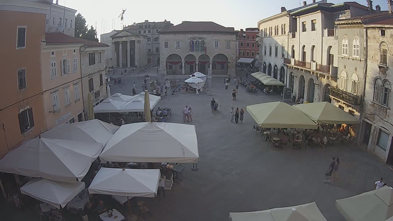 A town square with several outdoor cafes under shade umbrellas, surrounded by buildings in the late afternoon sun.