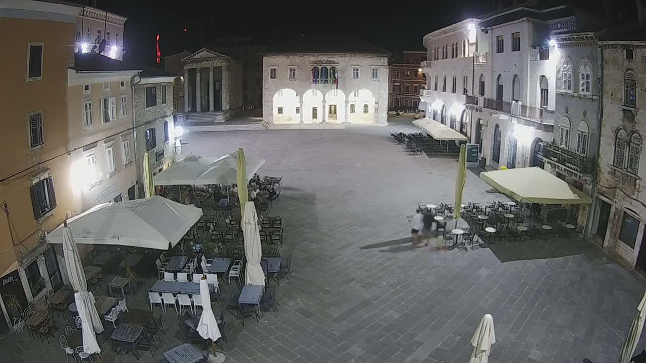 A nighttime, high-angle view of a town square with several outdoor cafes featuring tables, chairs, and umbrellas under bright artificial lighting.