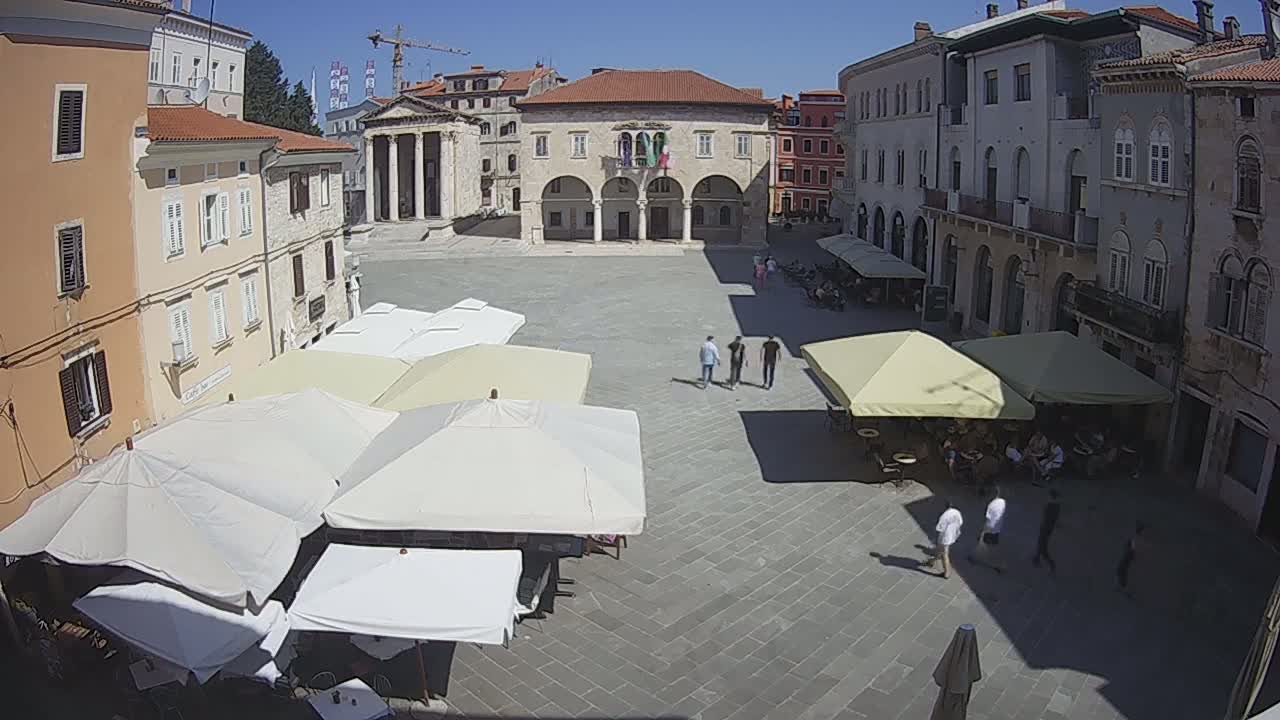 A sunny town square with several outdoor cafes under large umbrellas, surrounded by historic buildings.