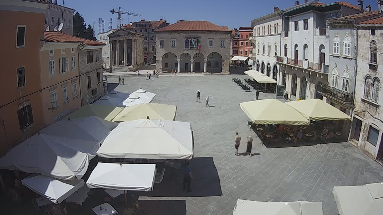 A sunny town square with several people walking around and outdoor seating under patio umbrellas.