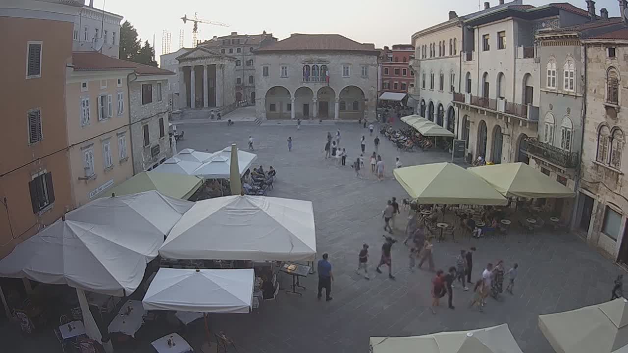 A town square with several people walking around and sitting under outdoor umbrellas on a partly cloudy day.