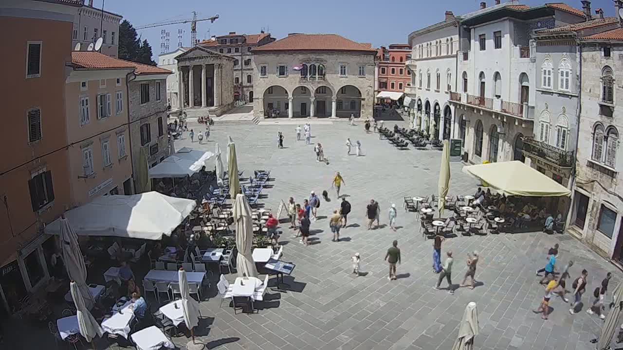 A sunny day reveals a large town square filled with people and outdoor cafe seating, surrounded by historic European-style buildings.