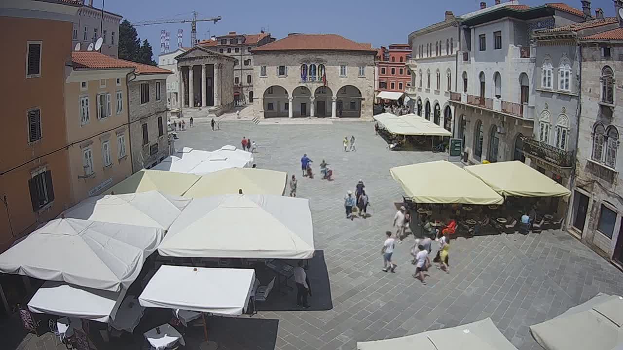 A town square with several outdoor cafes under shade umbrellas, surrounded by old buildings on a sunny day.