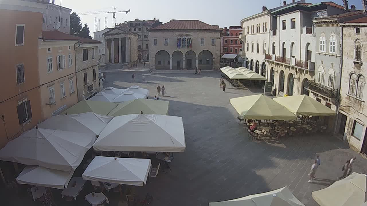 A town square with several outdoor cafes under umbrellas on a sunny day, surrounded by historic buildings.