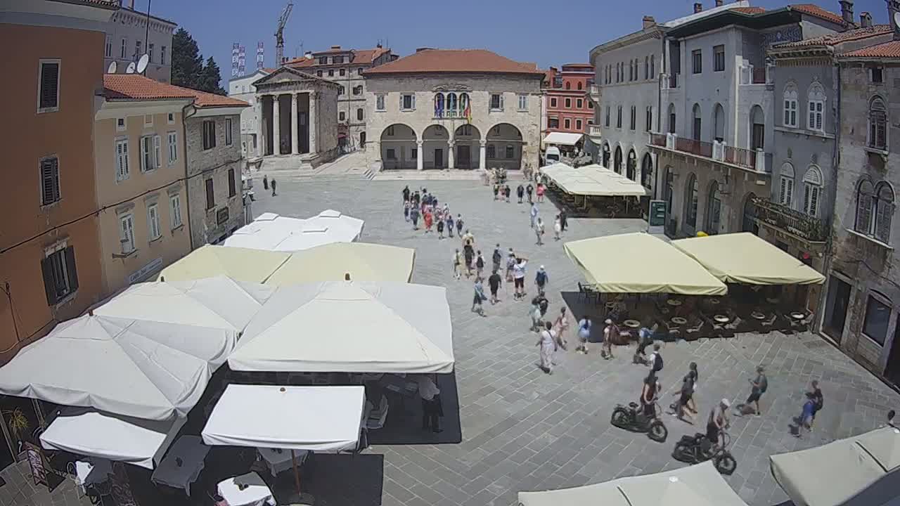 A sunny day in a town square with many people walking around, several outdoor cafe umbrellas, and stone buildings surrounding the square.