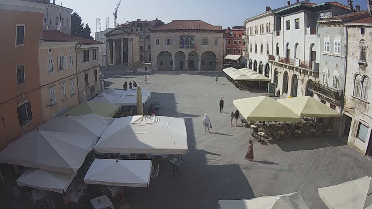 A sunny day in a large, paved European town square with several outdoor cafes featuring white and beige umbrellas and people walking around.