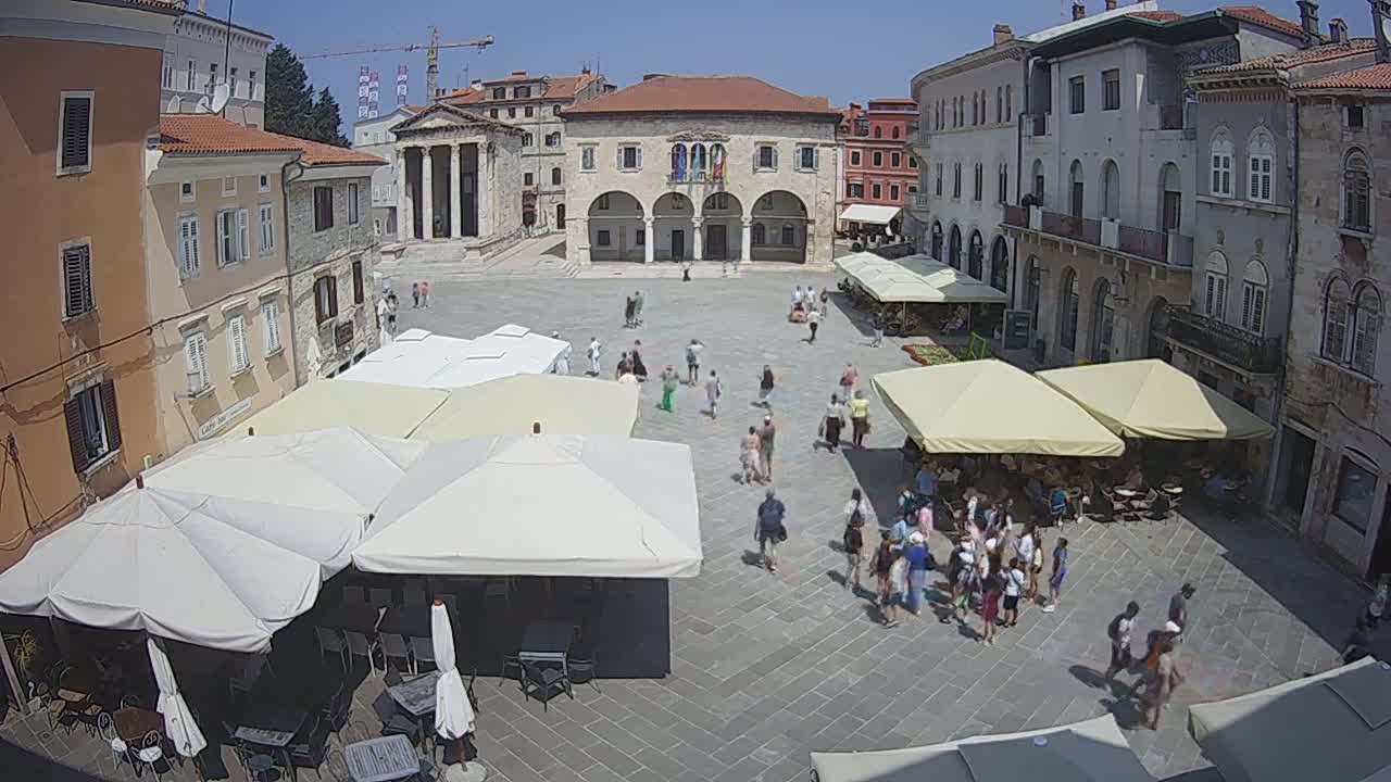 A sunny day in a large, stone-paved town square with numerous people strolling, and several outdoor cafes with shaded seating areas under umbrellas, surrounding a large building.
