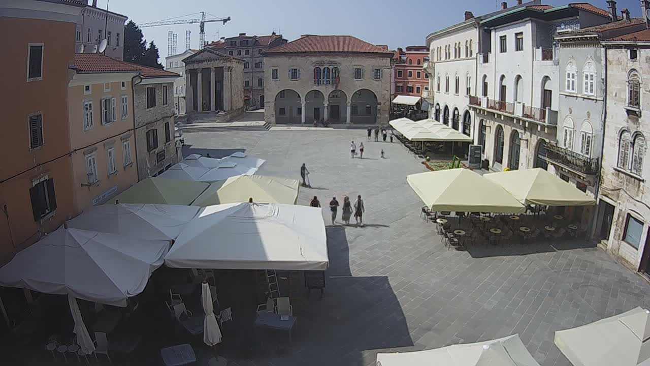 A sunny town square with several outdoor cafes under shade umbrellas, surrounded by historic buildings.