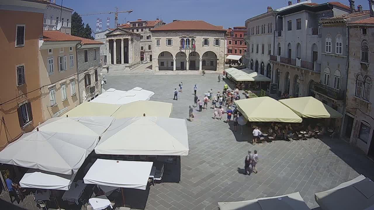 A sunny town square with several people milling about and numerous outdoor cafe umbrellas set up.