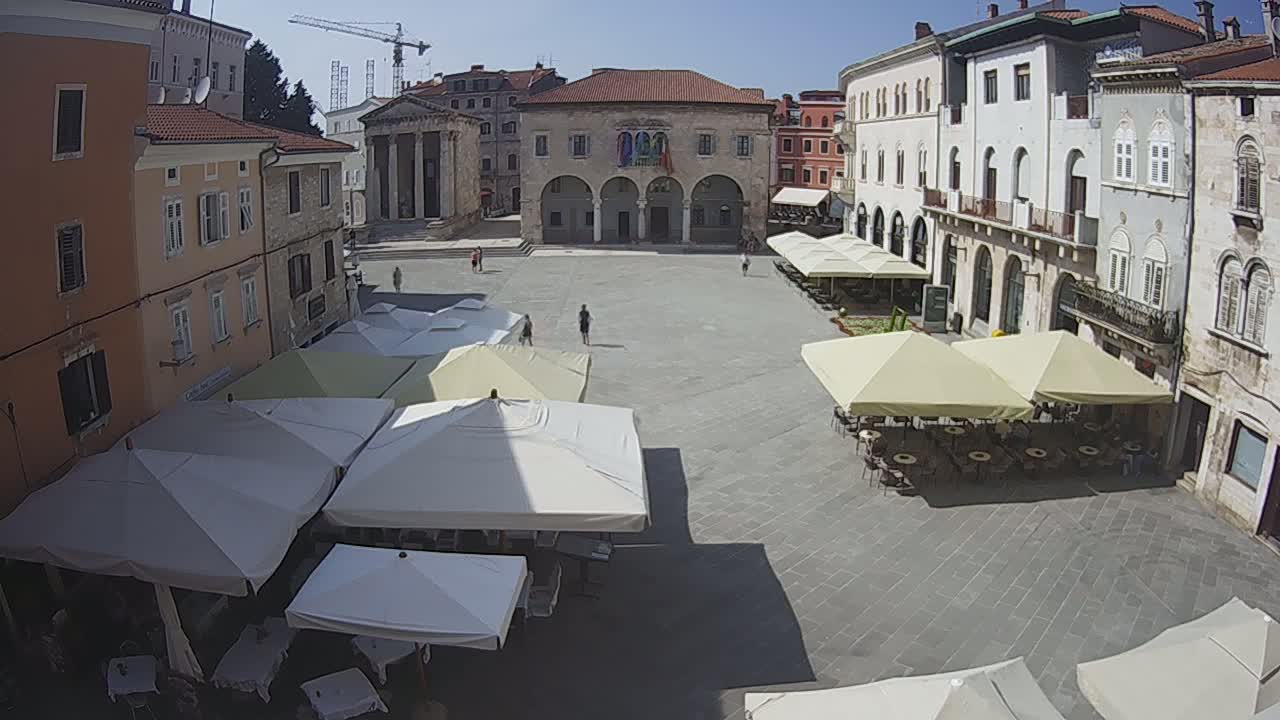 A large, paved town square is partially shaded by numerous outdoor cafe umbrellas on a sunny day, with several historic buildings surrounding it.