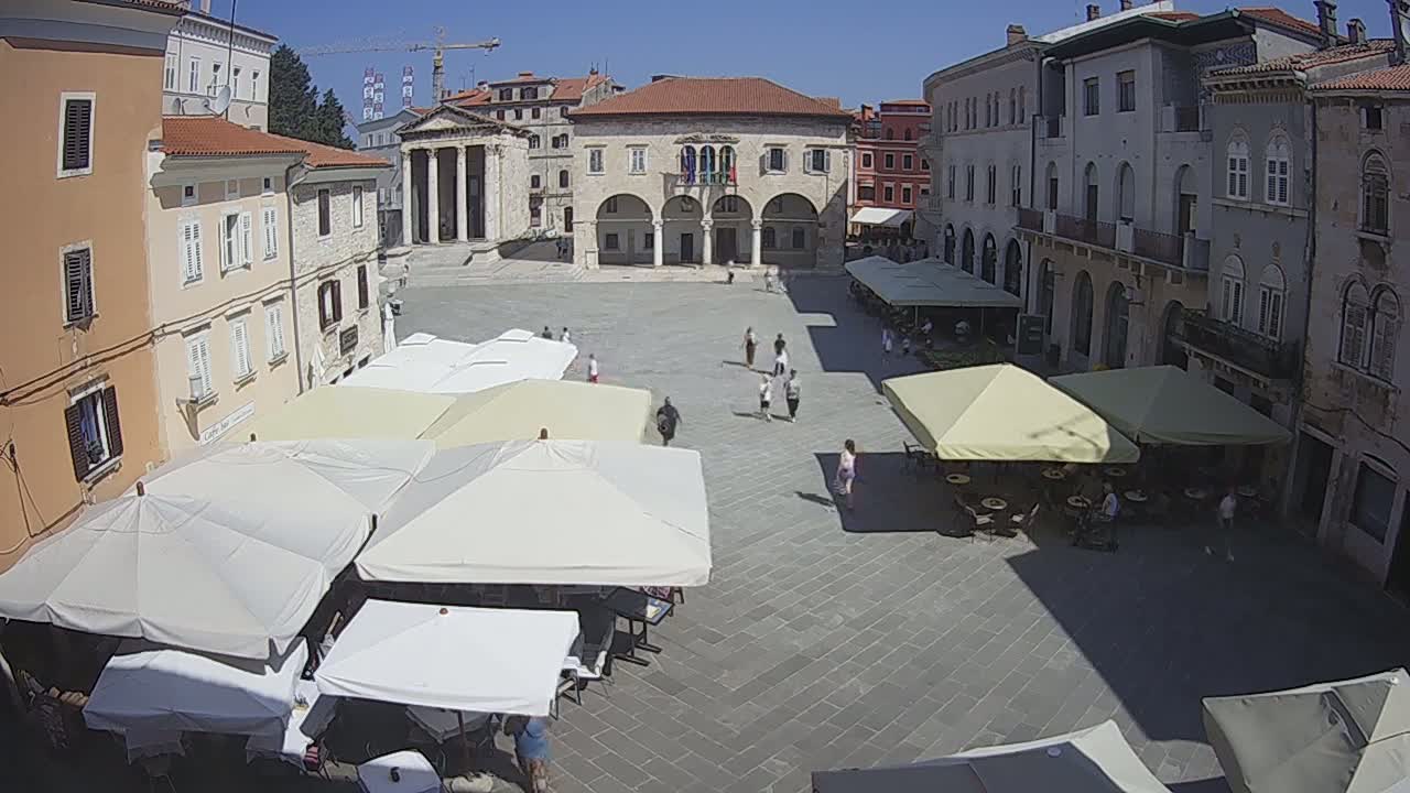 A sunny day in a European town square shows several people walking amongst numerous outdoor cafe umbrellas and historical buildings.