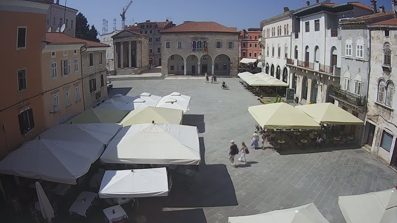 A sunny day in a European town square, showing several outdoor cafes with umbrellas set up around a large stone plaza and buildings.