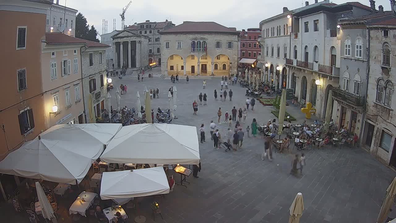 A sunny day in a European town square, showing several outdoor cafes with umbrellas set up around a large stone plaza and buildings.