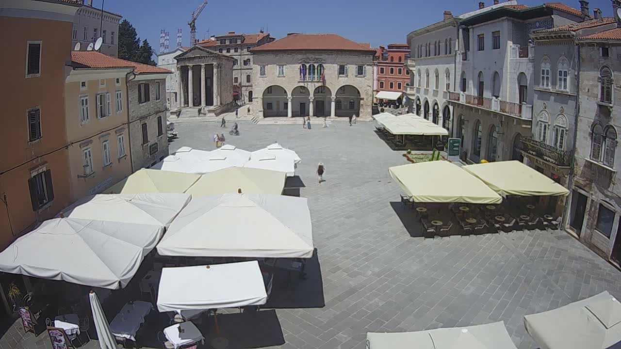 A sunny day reveals a large town square with several outdoor cafes under umbrellas, surrounded by historic buildings.