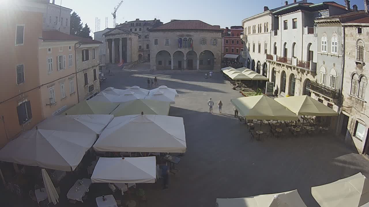 A sunny, paved town square is filled with several outdoor cafe umbrellas and a few pedestrians, surrounded by historic buildings.