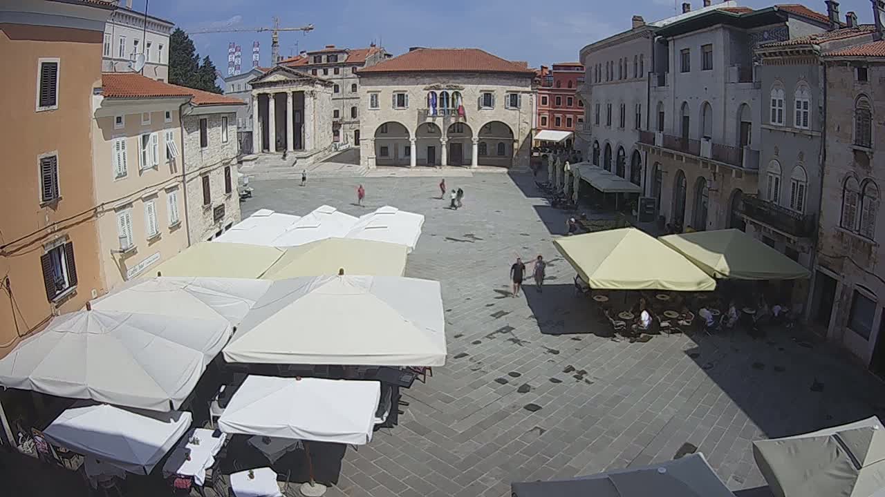 A town square with several outdoor cafe umbrellas and a large building in the background on a sunny day.