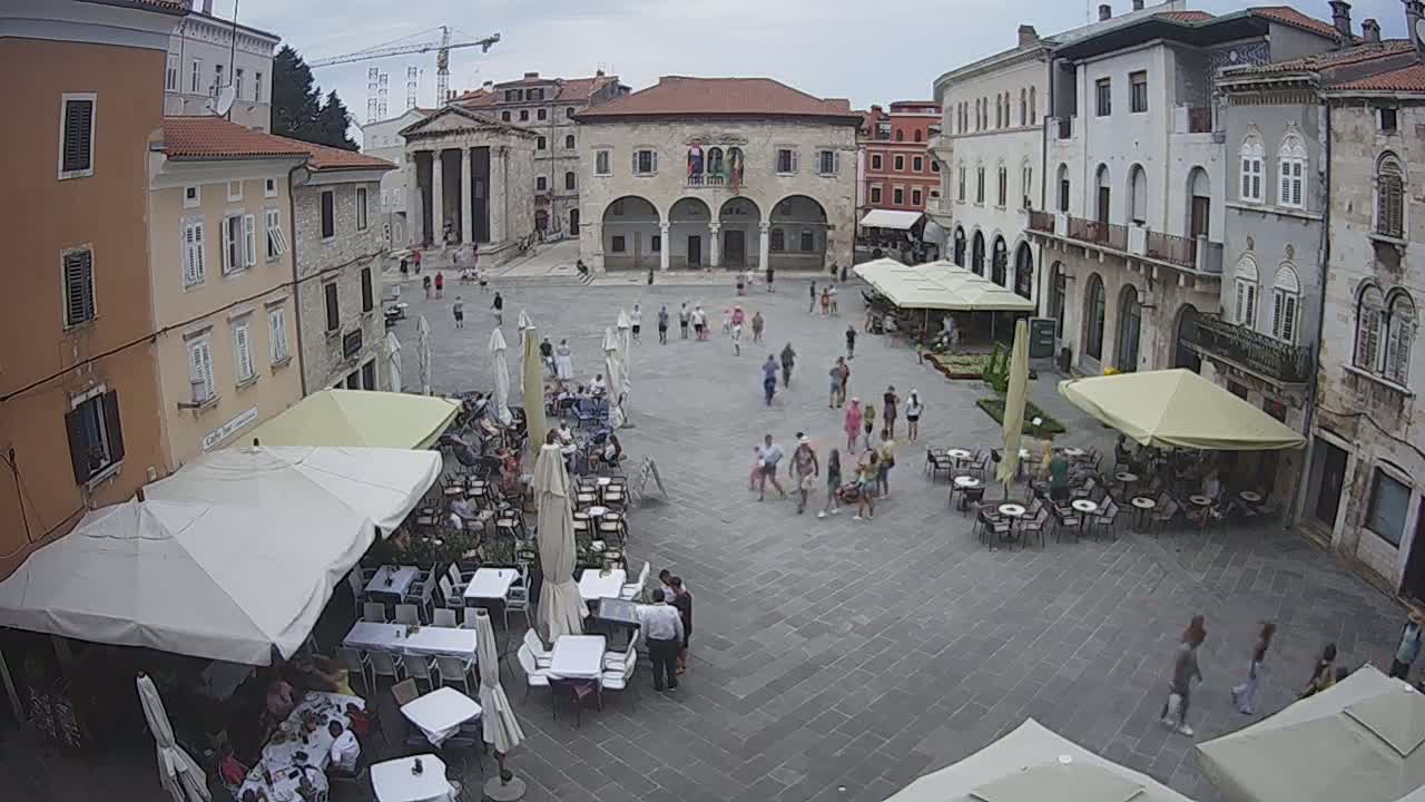 A town square with several outdoor cafes under umbrellas, surrounded by buildings, on an overcast day.