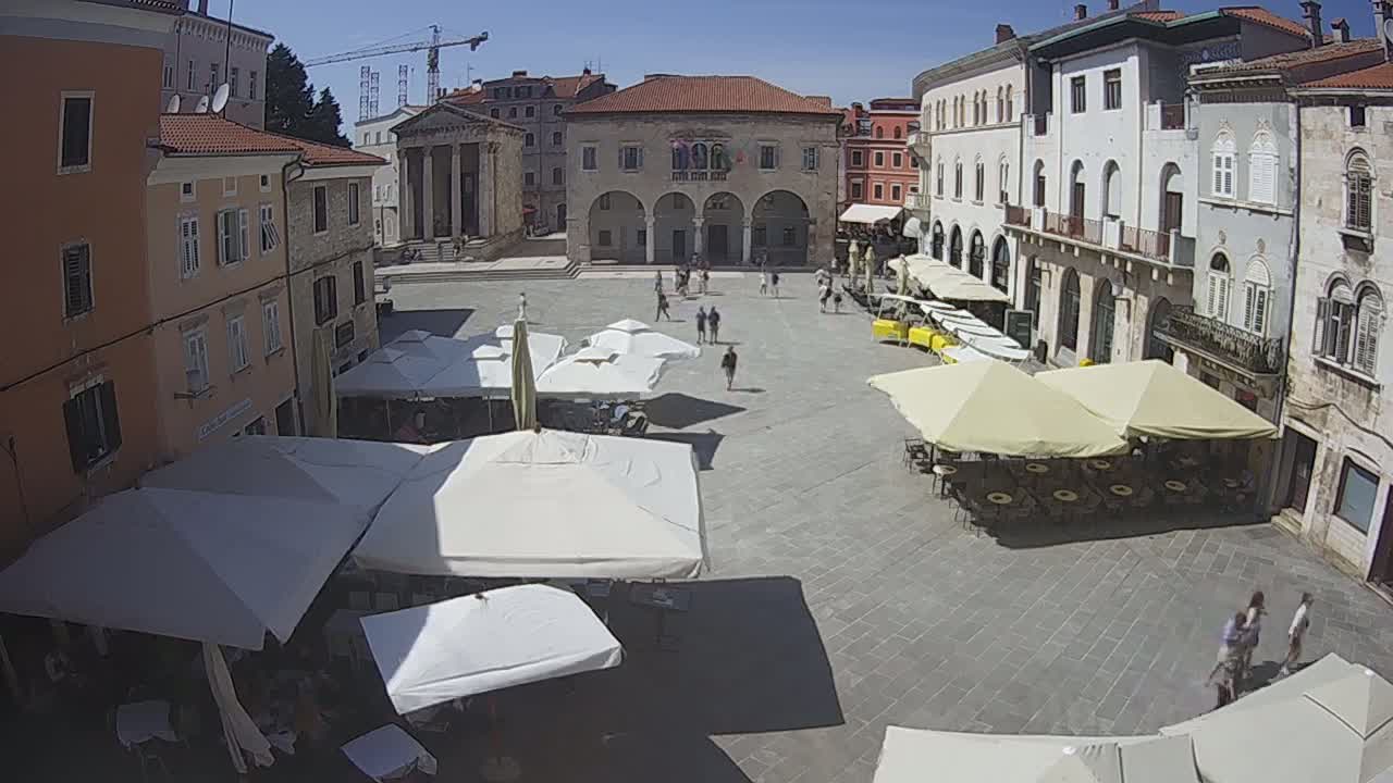 A sunny outdoor square in a European town features several cafe umbrellas under a clear blue sky, and several people are walking around the plaza.