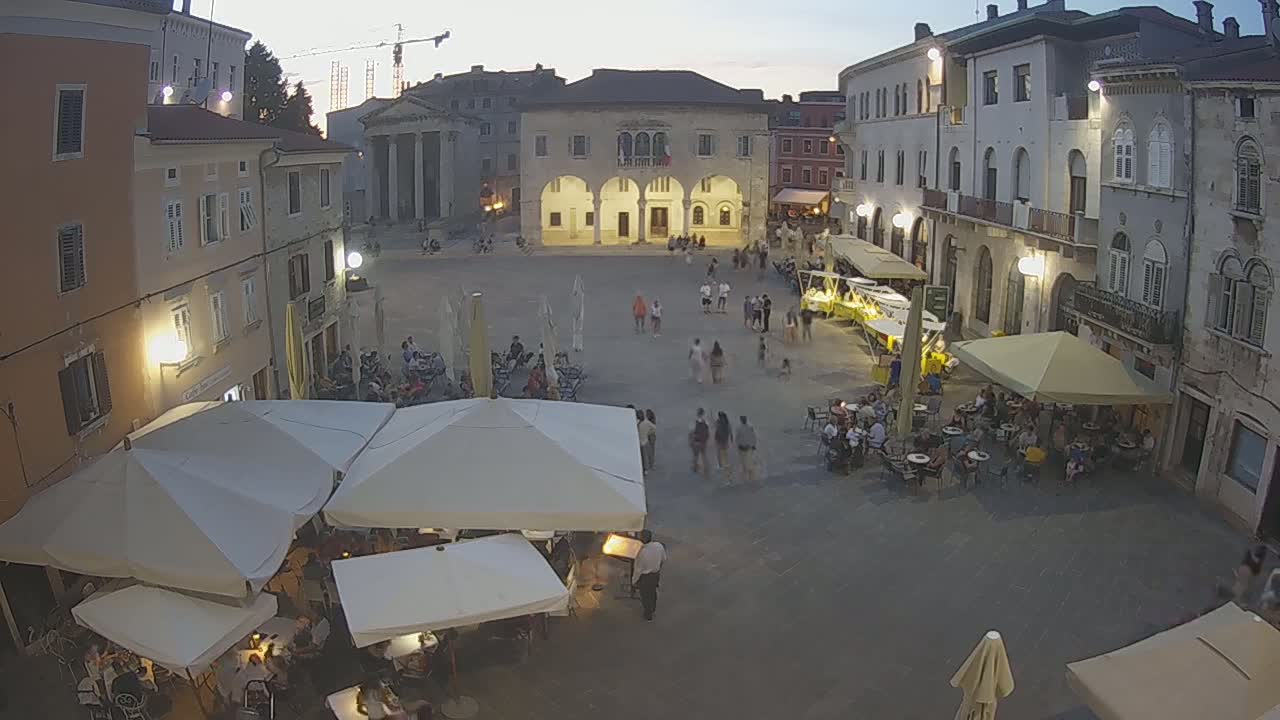 A town square at dusk, filled with people sitting at outdoor cafes under white umbrellas, surrounded by historic buildings.